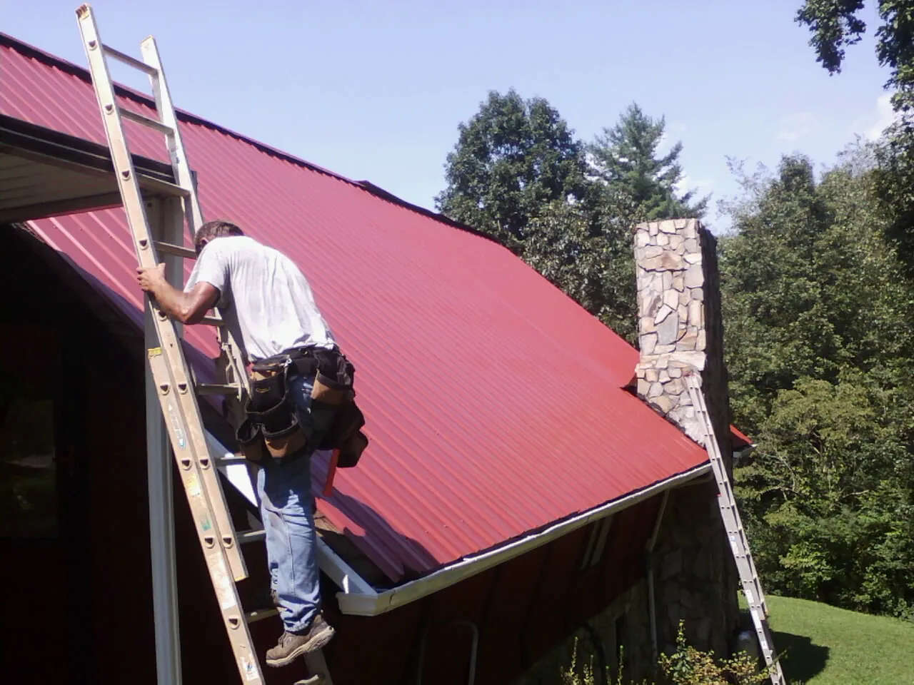 Roofer on a ladder beside a red metal roof and stone chimney — Soffit & Fascia Repair by Roofing Brothers in East Quogue
